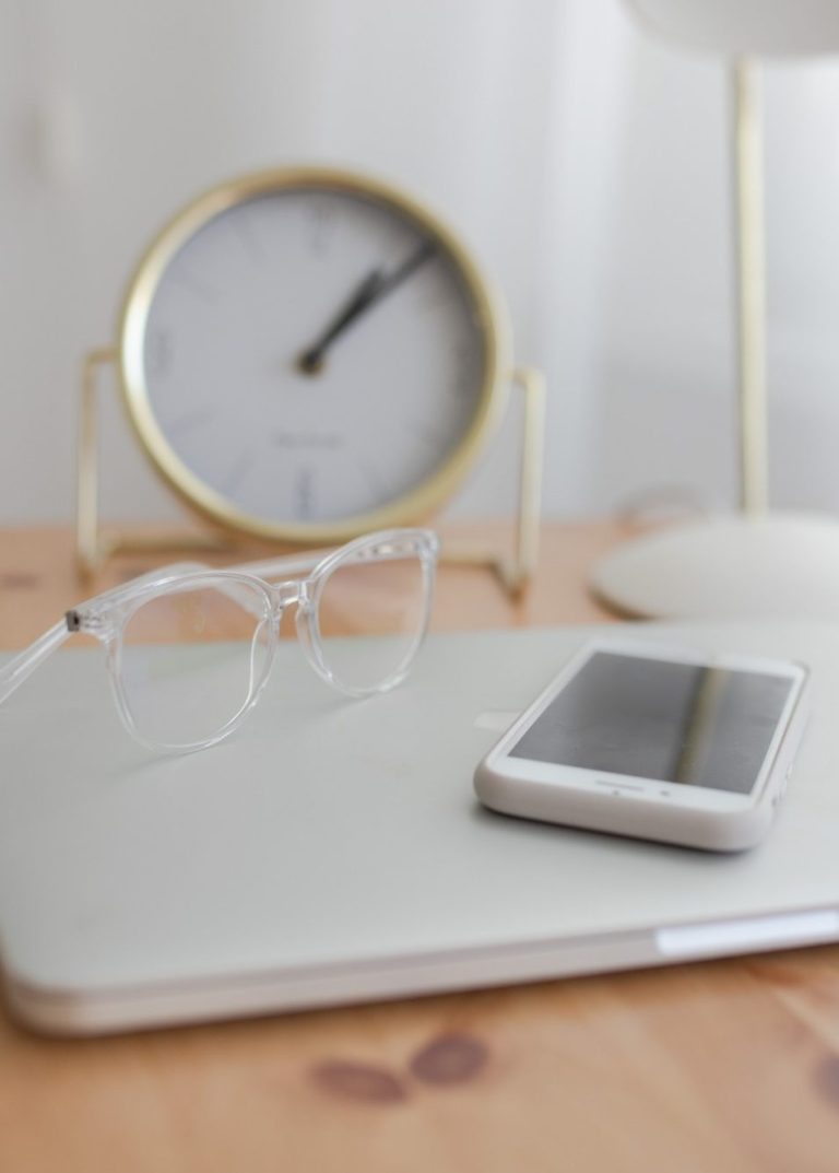 Une paire de lunettes et un téléphone portable sont posés sur un ordinateur. Dans le  fond de la photo une horloge avec un parement doré..