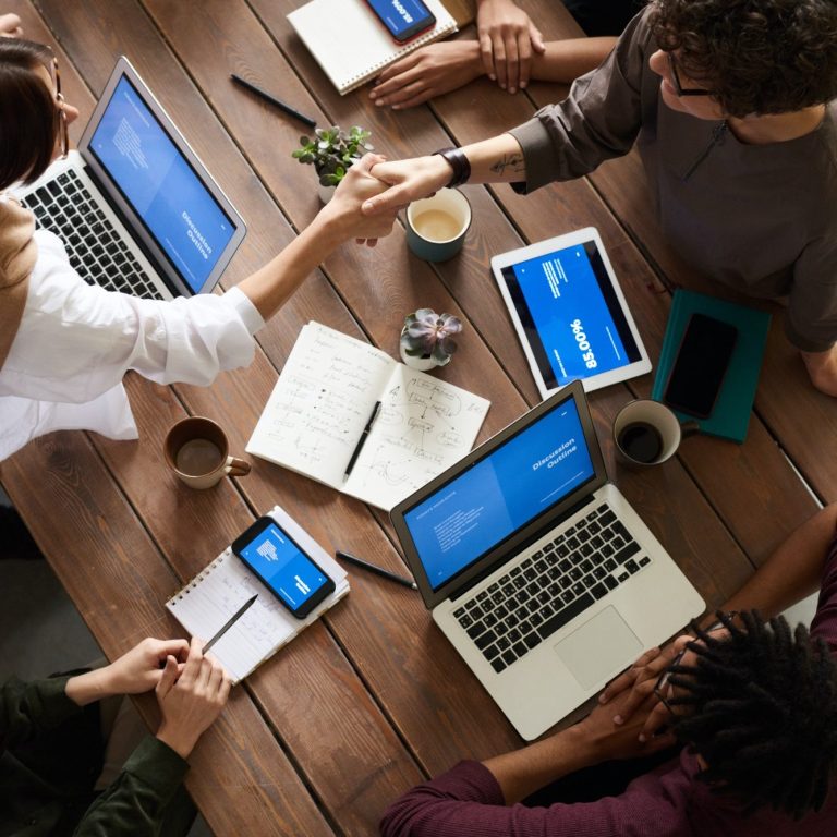 On voit des ordinateurs, tablette et téléphone de personnes qui travaillent sur le même bureau dont deux qui se serrent la main.